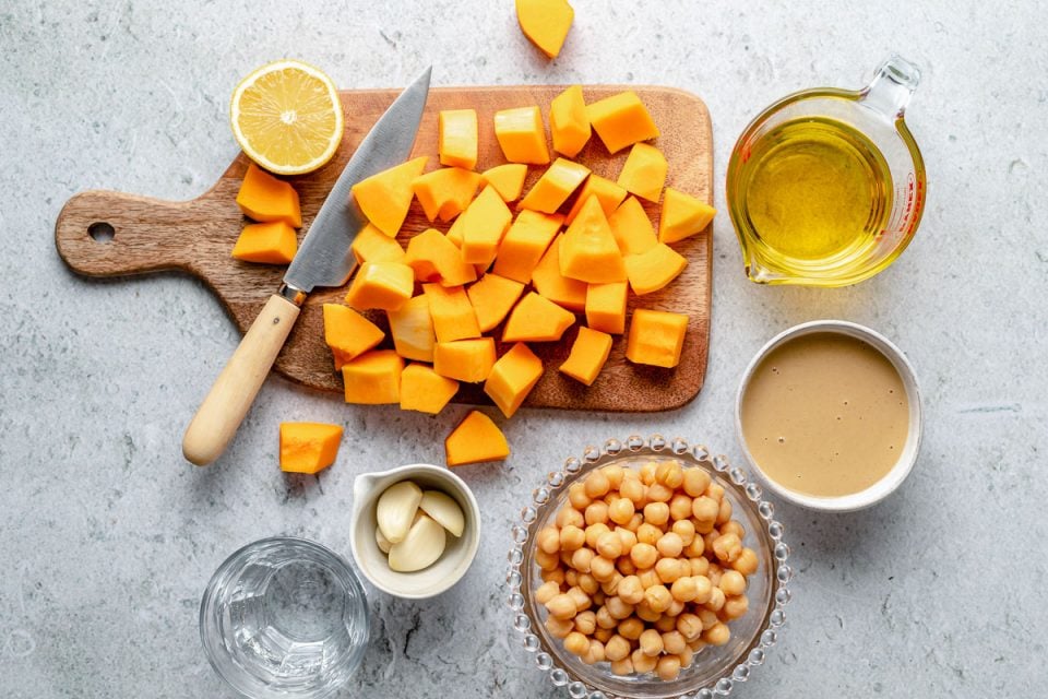 Overhead image of ingredients for butternut squash hummus including half of a lemon on a wood board, cubed butternut squash on a wood cutting board with a knife, olive oil in a glass measuring cup, tahini in a white bowl, chickpeas in a glass bowl, garlic cloves in a white measuring cup, and a glass of water.