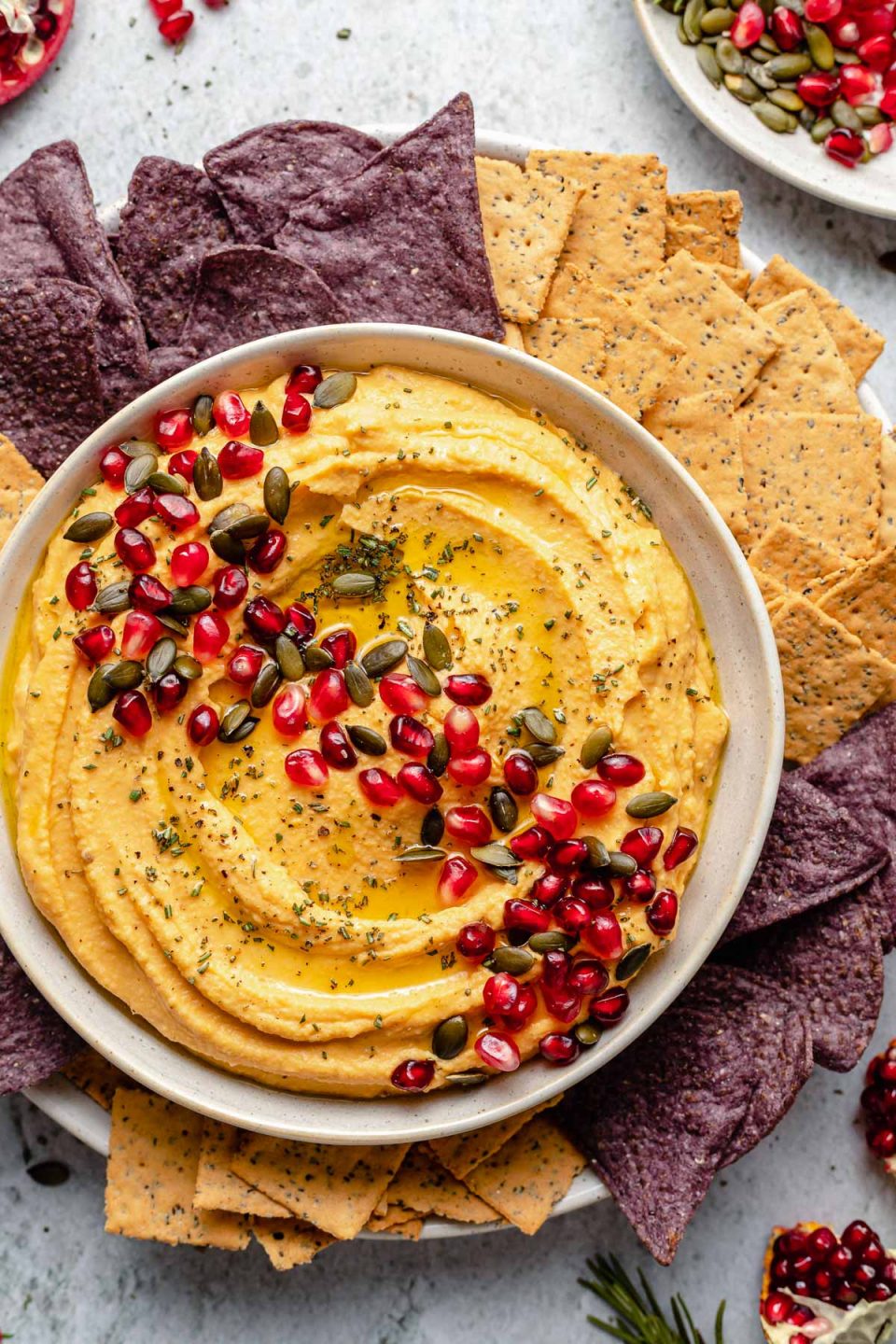 Overhead image of butternut squash hummus in a white serving bowl topped with pumpkin seeds, pomegranate arils, herbs, and olive oil. A variety of chips and crackers surround the bowl of hummus.