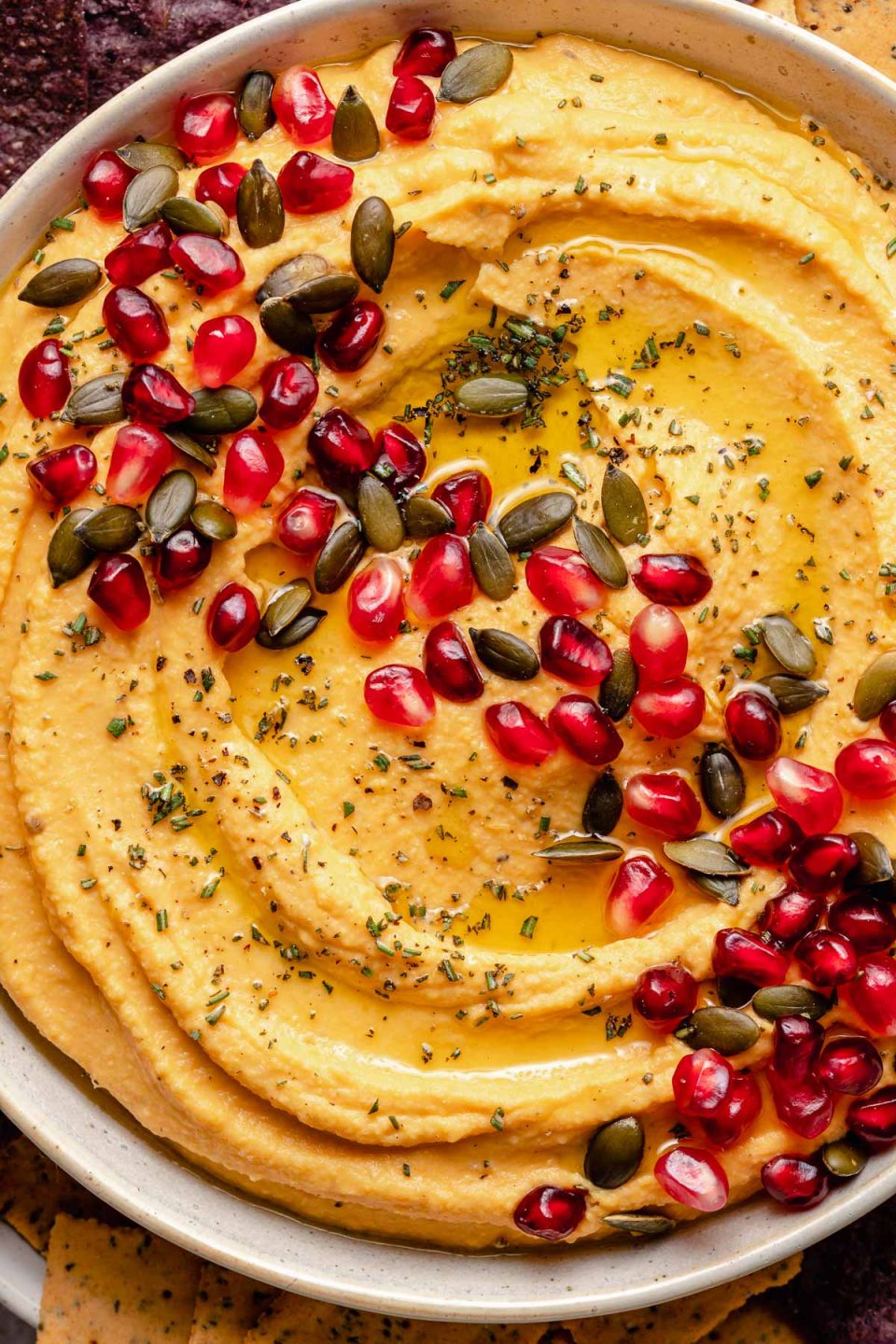 Close up overhead image of butternut squash hummus in a white bowl topped with pumpkin seeds, pomegranate arils, herbs, and olive oil.