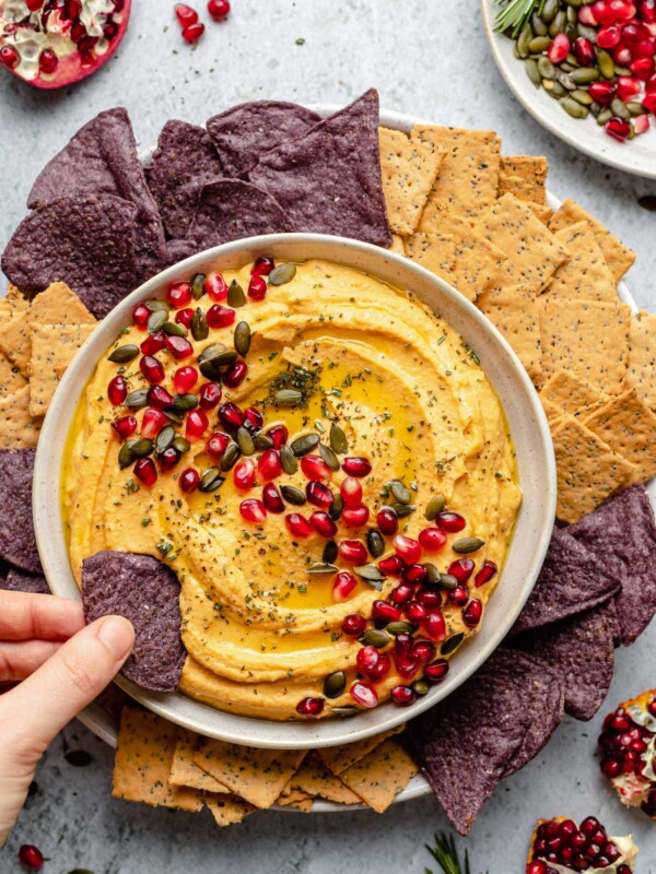 Overhead image of a hand dipping a blue corn tortilla chip into a bowl of butternut squash hummus. The hummus is in a white bowl topped with pumpkin seeds, pomegranate arils, and herbs. There are chips and crackers surrounding the bowl of hummus. There are sections of pomegranates in the corners of the image for garnish.