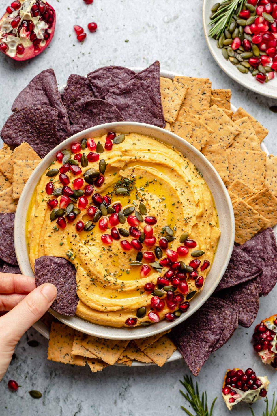 Overhead image of a hand dipping a blue corn tortilla chip into a bowl of butternut squash hummus. The hummus is in a white bowl topped with pumpkin seeds, pomegranate arils, and herbs. There are chips and crackers surrounding the bowl of hummus. There are sections of pomegranates in the corners of the image for garnish.