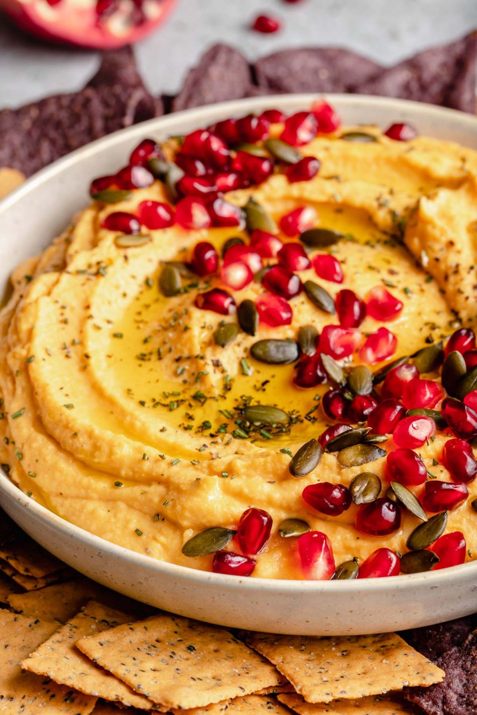 Close up of butternut squash hummus in a white serving bowl topped with pumpkin seeds, pomegranate arils, herbs, and olive oil. Next to the bowl are a variety of chips and crackers.