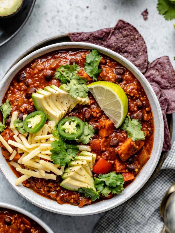 Sweet Potato Quinoa Chili shown in 2 soup bowls, topped with avocado, jalapeno, shredded vegan cheese, lime wedges & cilantro leaves. The bowls sit atop a light blue surface, next to a checkered blue & white linen, soup spoons, garnishes (sliced jalapeno, lime wedges, tortilla chips, etc.).