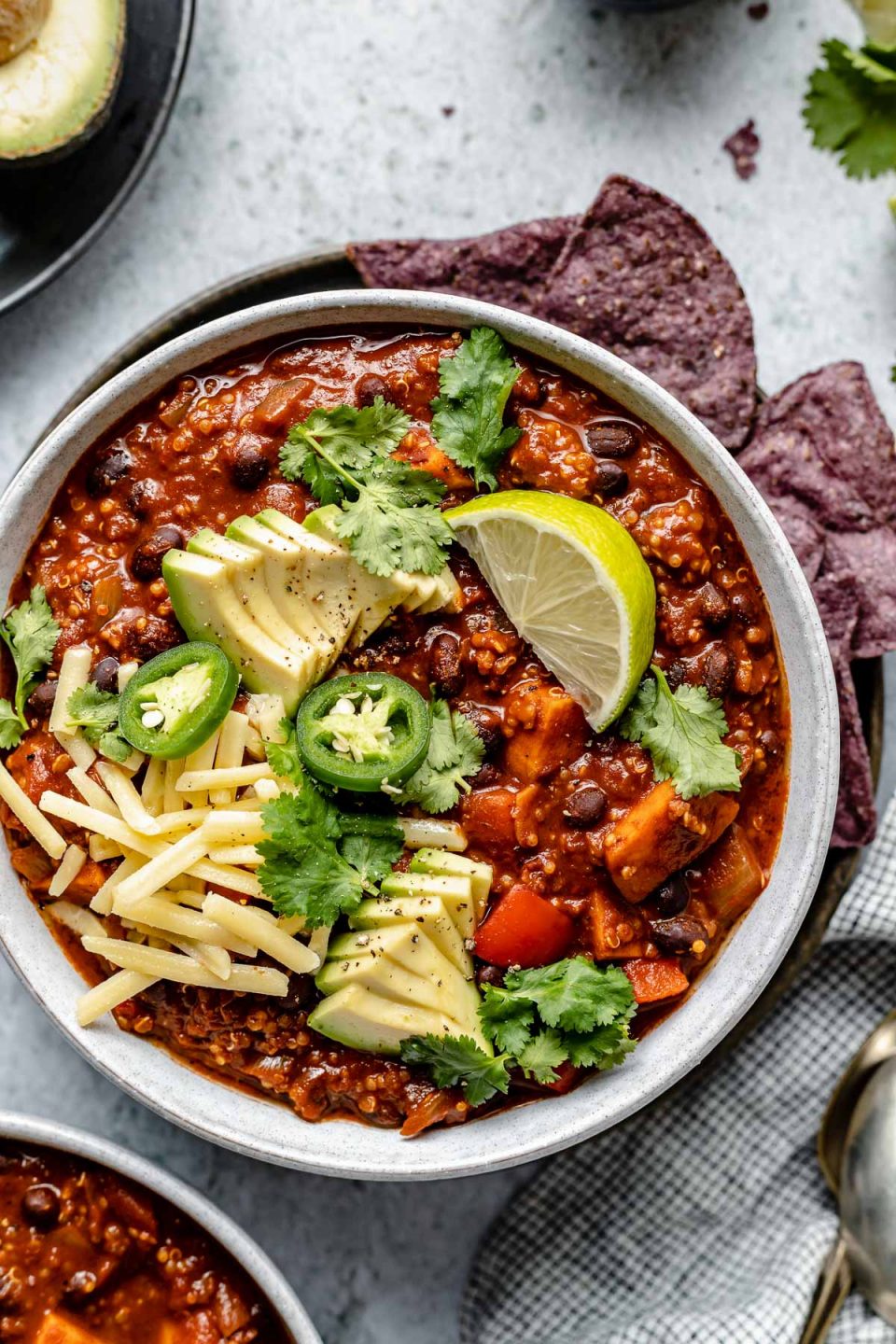 Sweet Potato Quinoa Chili shown in 2 soup bowls, topped with avocado, jalapeno, shredded vegan cheese, lime wedges & cilantro leaves. The bowls sit atop a light blue surface, next to a checkered blue & white linen, soup spoons, garnishes (sliced jalapeno, lime wedges, tortilla chips, etc.).