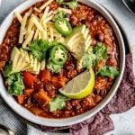 Sweet Potato Quinoa Chili shown in 2 soup bowls, topped with avocado, jalapeno, shredded vegan cheese, lime wedges & cilantro leaves. The bowls sit atop a light blue surface, next to a checkered blue & white linen, soup spoons, garnishes (sliced jalapeno, lime wedges, tortilla chips, etc.).