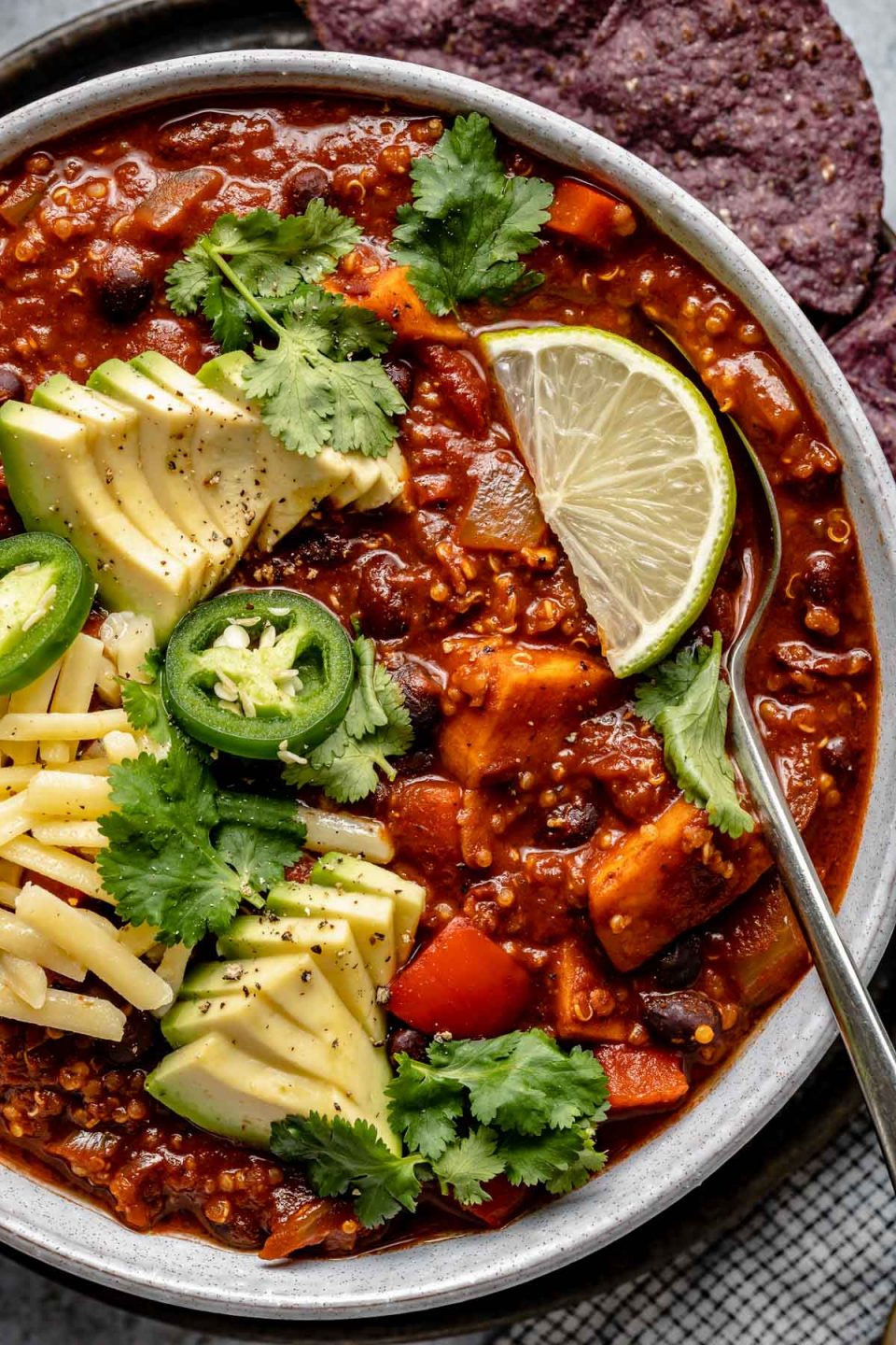 Close up overhead of Sweet Potato Quinoa Chili shown in a soup bowl, topped with avocado, jalapeno, shredded vegan cheese, lime wedges & cilantro leaves. A spoon is nestled in the bowl of chili.