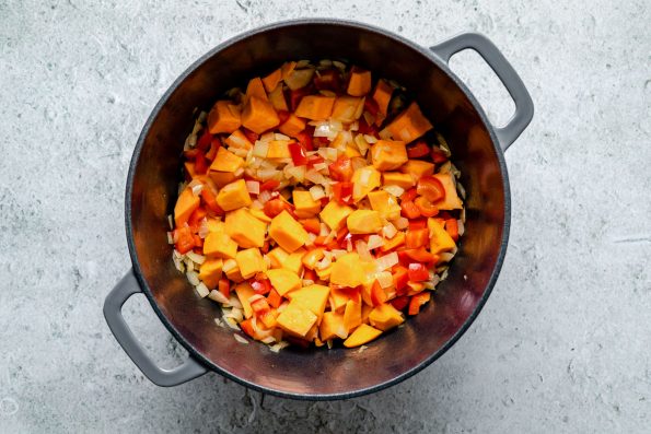 How to make Sweet Potato Quinoa Chili - Step 1: Onions, bell pepper, & sweet potato cooking in a large gray Dutch oven, which sits atop a light blue surface.