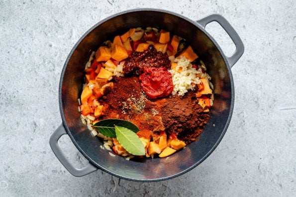 How to make Sweet Potato Quinoa Chili - Step 2: Spices (chipotle pepper, tomato paste, garlic, cocoa powder, chili powder, paprika, bay leaves) added to chili veggies in a large gray Dutch oven, which sits atop a light blue surface.