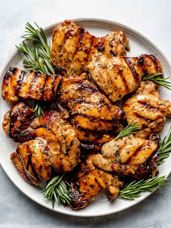 Grilled brown sugar bourbon chicken thighs shown on a white speckled plate atop a light blue surface. The chicken is garnished with fresh sprigs of rosemary.