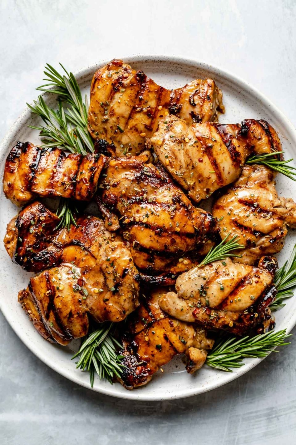 Grilled brown sugar bourbon chicken thighs shown on a white speckled plate atop a light blue surface. The chicken is garnished with fresh sprigs of rosemary.