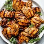 Grilled brown sugar bourbon chicken thighs shown on a white speckled plate atop a light blue surface. The chicken is garnished with fresh sprigs of rosemary.