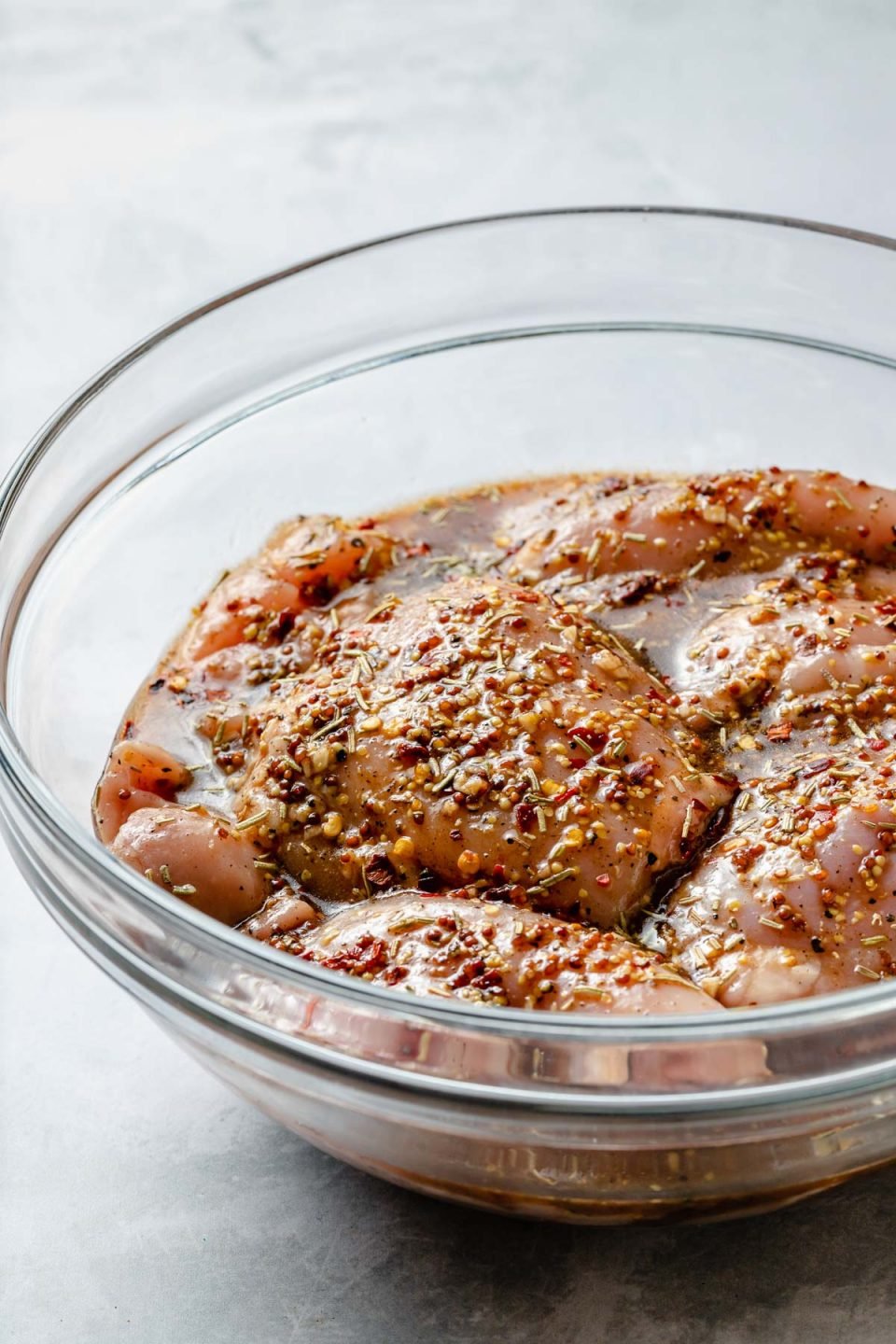 Side angle of chicken thighs in a large glass mixing bowl, marinating in Brown Sugar Bourbon marinade. The bowl sits atop a light blue surface.