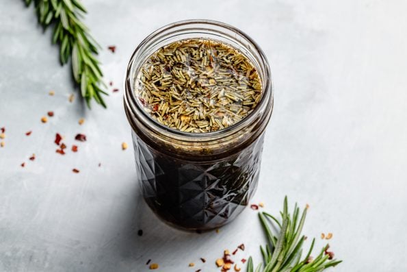 Brown Sugar Bourbon marinade shown in a small ball jar, sitting atop a light blue surface with a fresh sprig of rosemary & crushed red chili pepper flakes on the surface in the foreground & in the background.