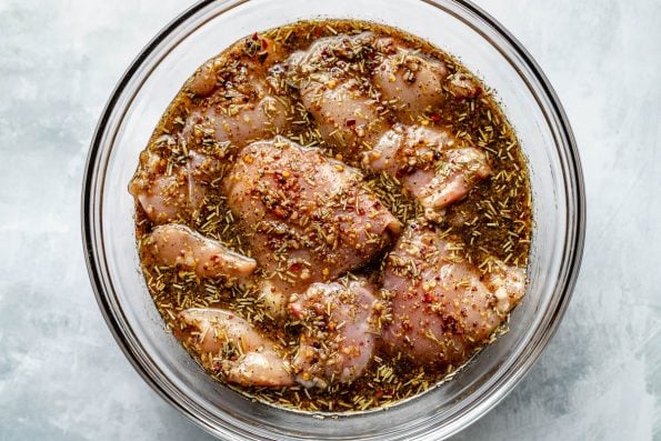 Chicken thighs in a large glass mixing bowl, marinating in Brown Sugar Bourbon marinade. The bowl sits atop a light blue surface.