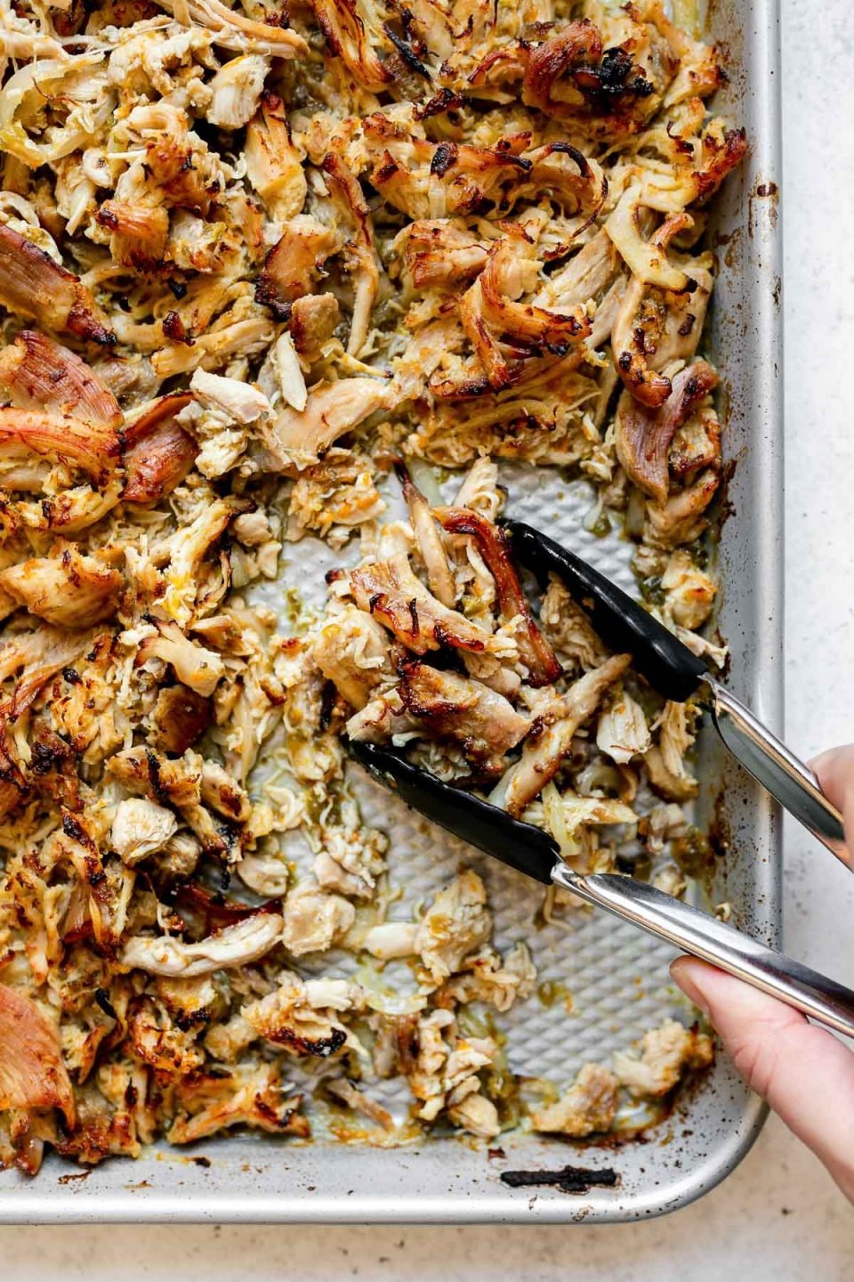 Shredded crispy chicken carnitas on a small silvery baking sheet, which sits atop a white surface. A woman's hand reaches into the frame with small tongs, scooping up some of the chicken carnitas.