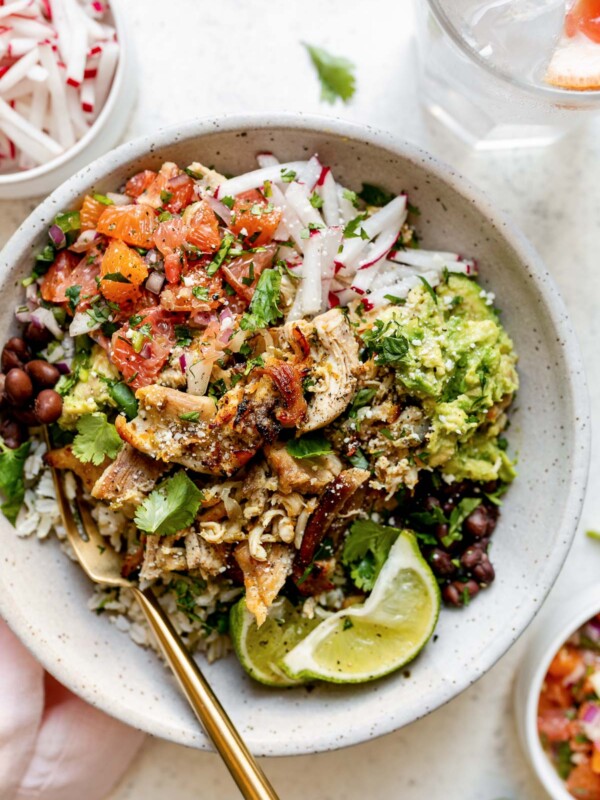 Chicken carnitas rice bowl, with rice, beans, guacamole, shredded radishes, winter citrus pico de gallo. The bowl sits atop a white surface, next to a pink linen napkin, small bowls of winter citrus pico de gallo & shredded radish, & a glass of sparkling water with lemon & grapefruit wedges in it.