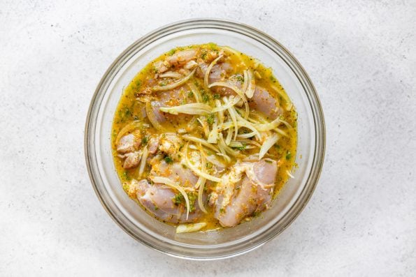 How to make chicken carnitas, Step 1: Chicken thighs & yellow onion marinating in citrus & jalapeno carnitas sauce in a large glass bowl, which sits atop a white surface.