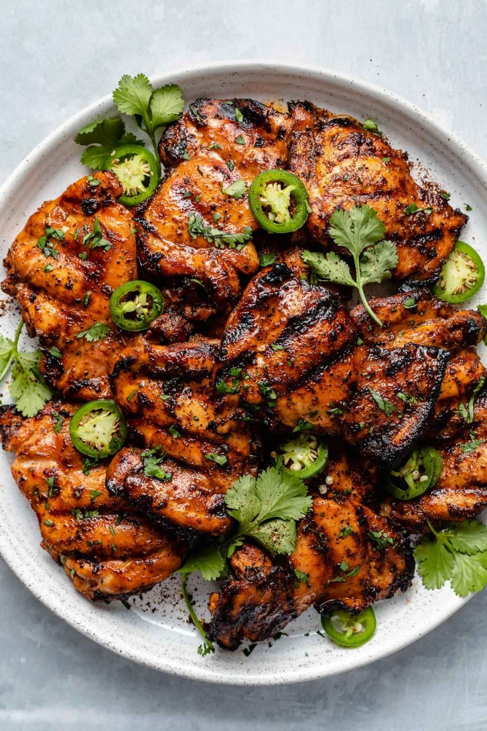 Grilled chicken thighs shown on a white speckled plate atop a light blue surface. The chicken is garnished with fresh cilantro leaves, sliced jalapeño, & ground black pepper.