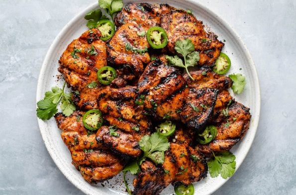 Grilled chili lime chicken thighs shown on a white speckled plate atop a light blue surface. The chicken is garnished with fresh cilantro leaves, sliced jalapeño, & ground black pepper.