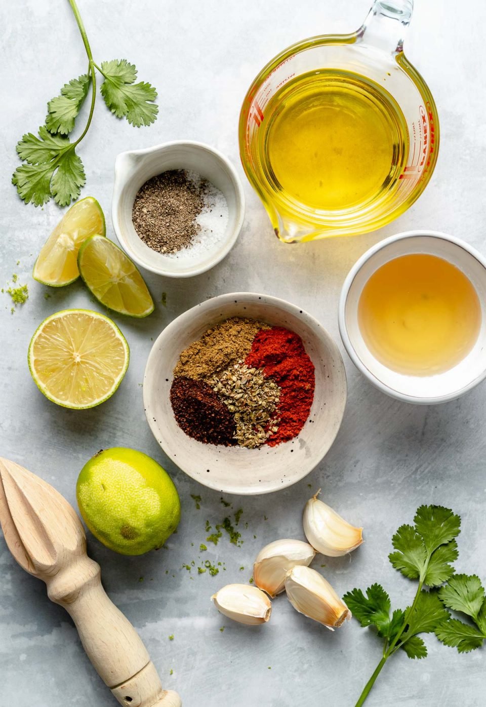 Ingredients are arranged on a light blue surface: olive oil, limes, garlic, agave, ancho chile powder, ground cumin, smoked paprika, dried oregano, salt, & pepper. A wooden citrus juicer & fresh cilantro leaves rest among the ingredients.