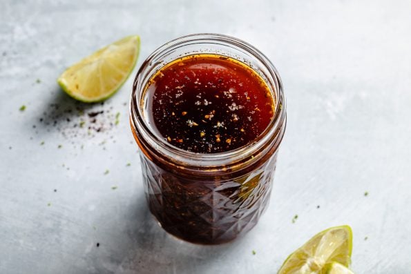 Chili Lime marinade shown in a small ball jar, sitting atop a light blue surface with lime wedges, lime zest, & ground black pepper on the surface in the foreground & in the background.