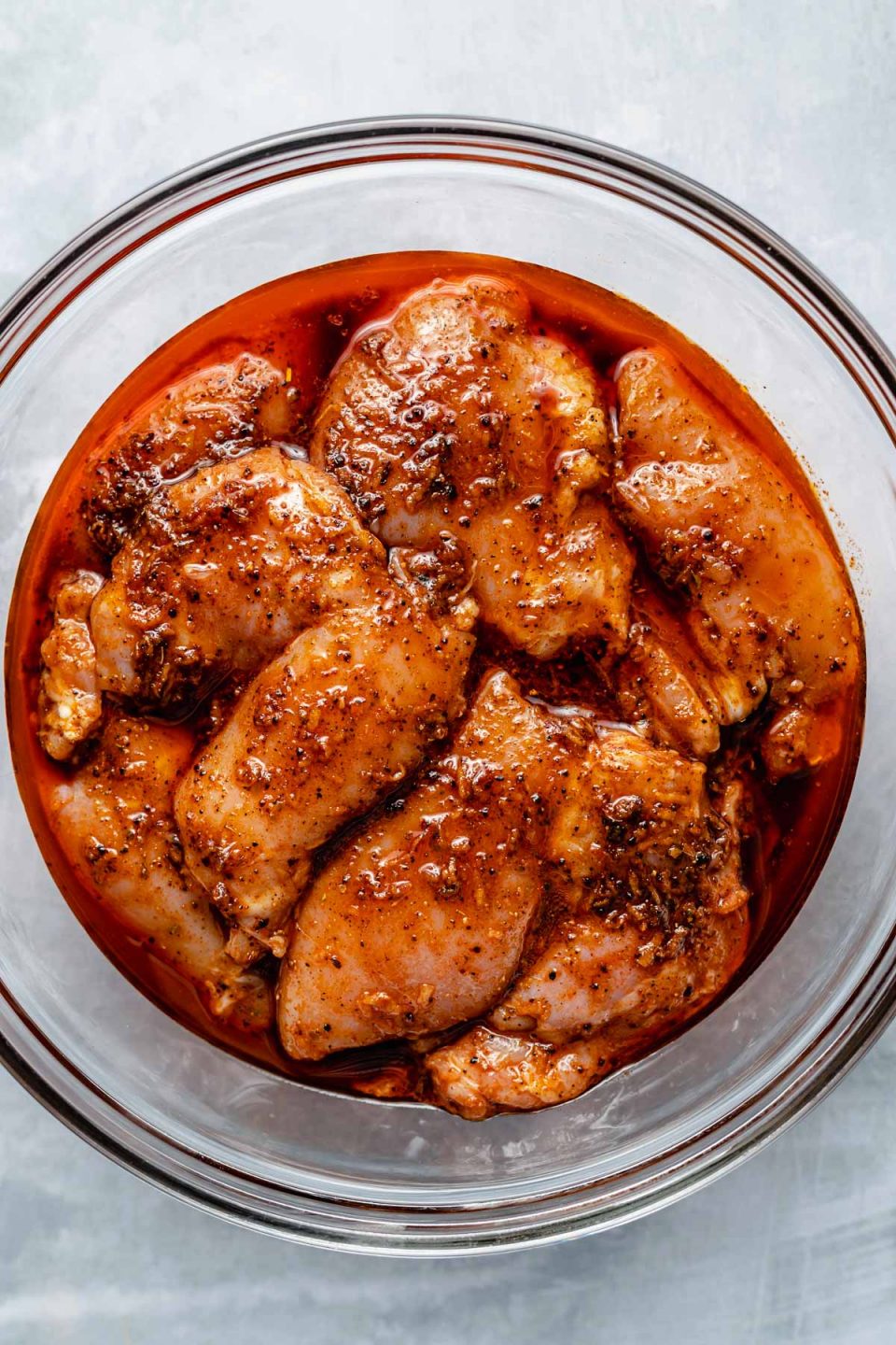 Chicken thighs in a large glass mixing bowl, marinating in Chili Lime marinade. The bowl sits atop a light blue surface.