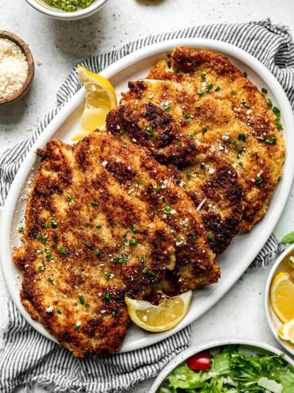 Parmesan crusted chicken cutlets on a large white platter, which sits atop a gray striped linen napkin on a white surface, surrounded by salad, a bowl of lemon wedges, a small bowl of pesto, & a small bowl of grated parmesan cheese.