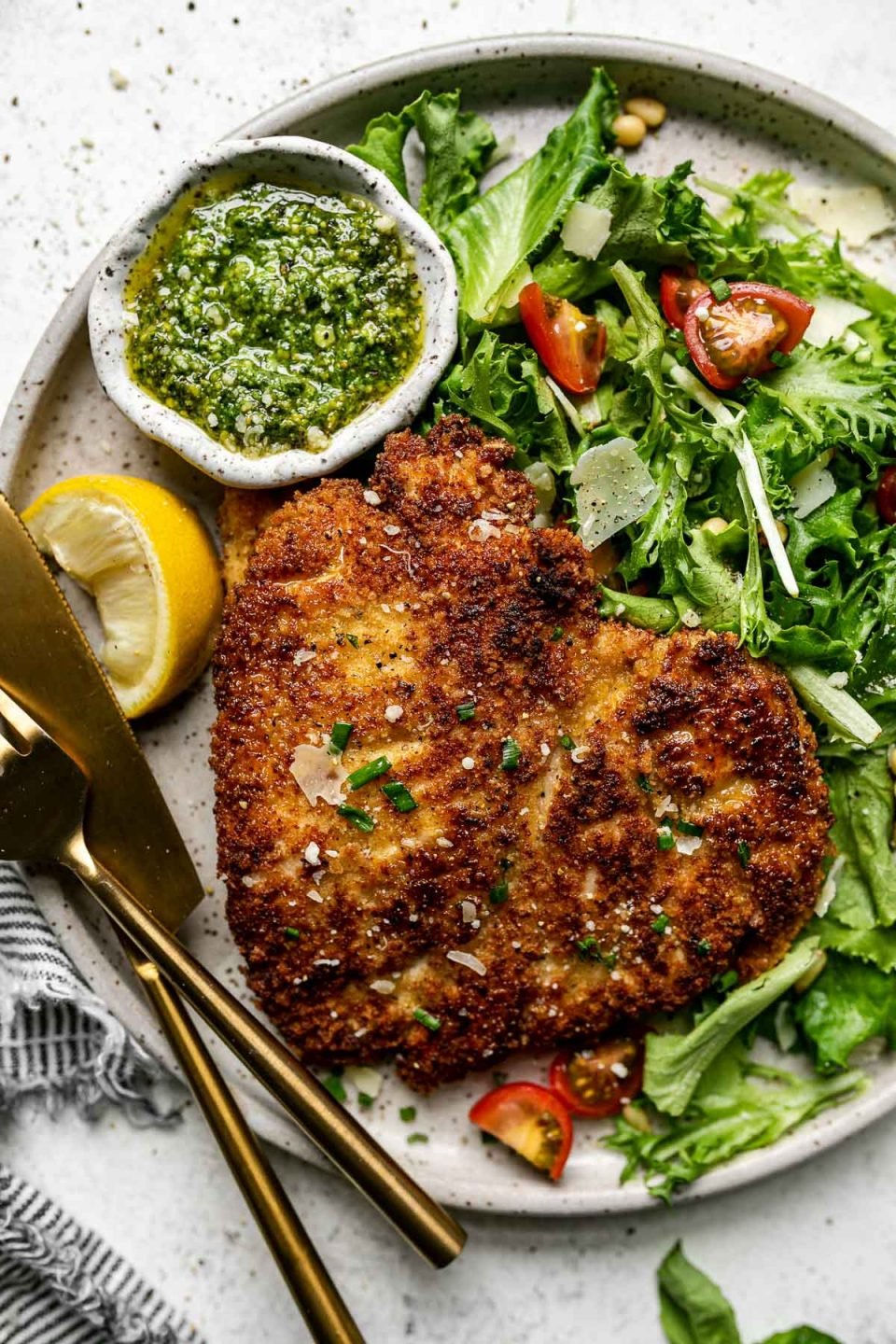 Parmsean chicken breast on a large plate with a green salad & a small bowl of pesto. The plate sits atop a white surface, next to a gray striped linen napkin.