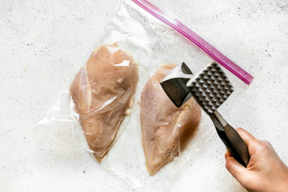 How to make parmesan crusted chicken, Step 1: Chicken breasts in a large plastic bag on a white surface. A woman's hand reaches into the frame with a meat mallet, pounding them flat.