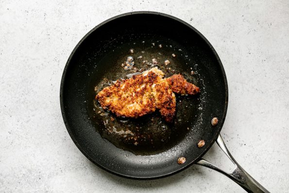 How to make parmesan crusted chicken, Step 4: Pan frying parmesan crusted chicken in a black nonstick skillet, which sits atop a white surface.