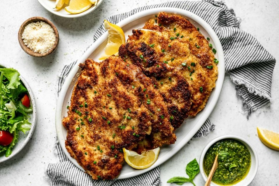 Parmesan crusted chicken cutlets on a large white platter, which sits atop a gray striped linen napkin on a white surface, surrounded by salad, a bowl of lemon wedges, a small bowl of pesto, & a small bowl of grated parmesan cheese.