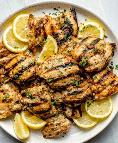 Grilled Greek chicken thighs shown on a white speckled plate atop a light blue surface. The chicken is garnished with fresh chopped herbs, lemon slices, & lemon wedges.
