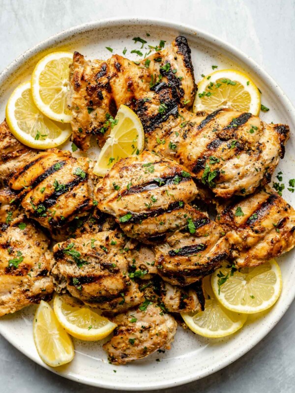 Grilled Greek chicken thighs shown on a white speckled plate atop a light blue surface. The chicken is garnished with fresh chopped herbs, lemon slices, & lemon wedges.