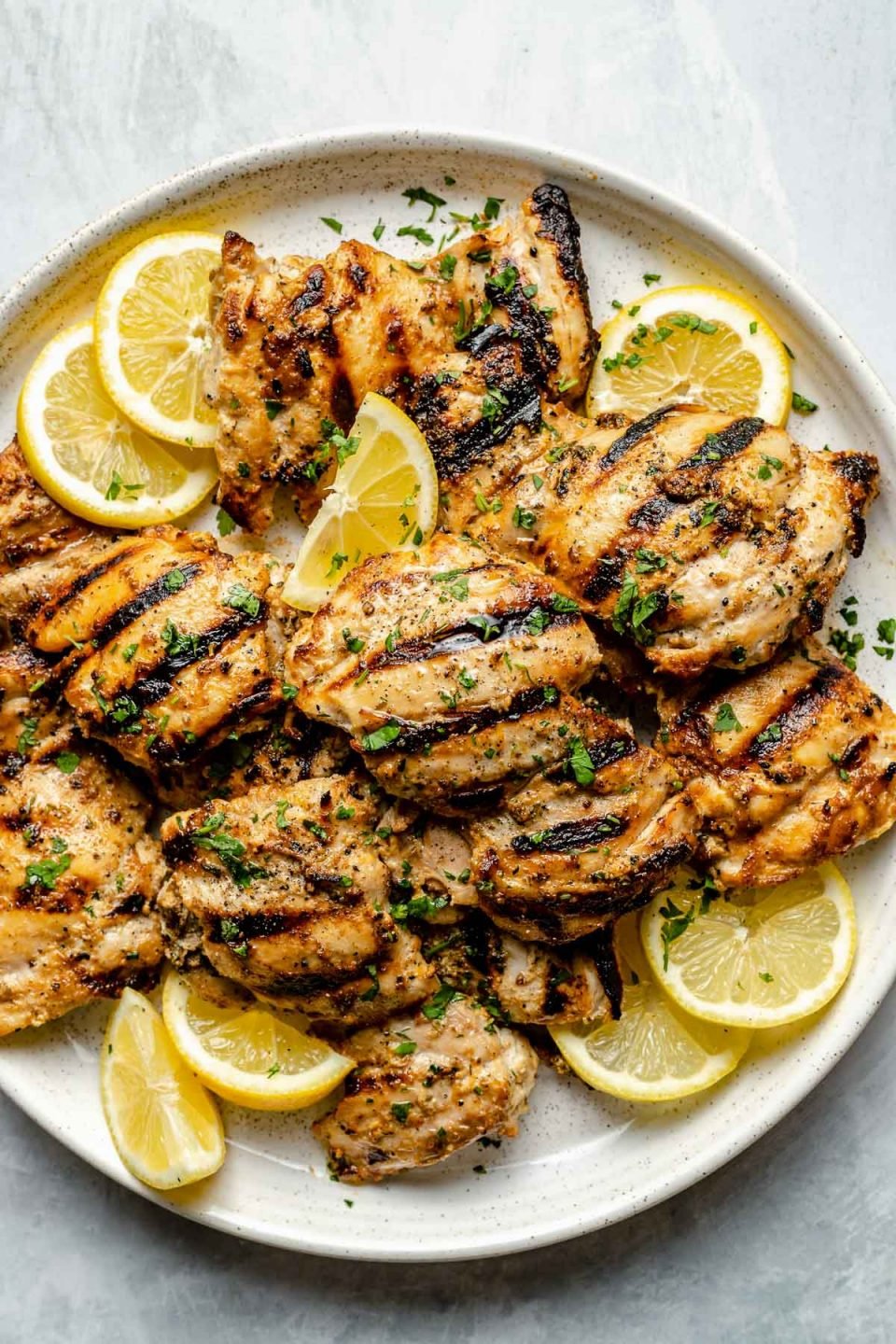 Grilled Greek chicken thighs shown on a white speckled plate atop a light blue surface. The chicken is garnished with fresh chopped herbs, lemon slices, & lemon wedges.