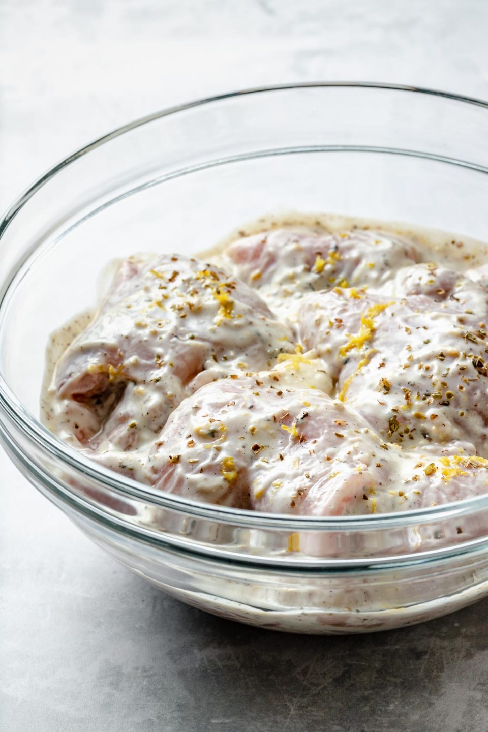 Side angle of chicken thighs in a large glass mixing bowl, marinating in Greek marinade. The bowl sits atop a light blue surface. Fresh lemon zest has been sprinkled on top.