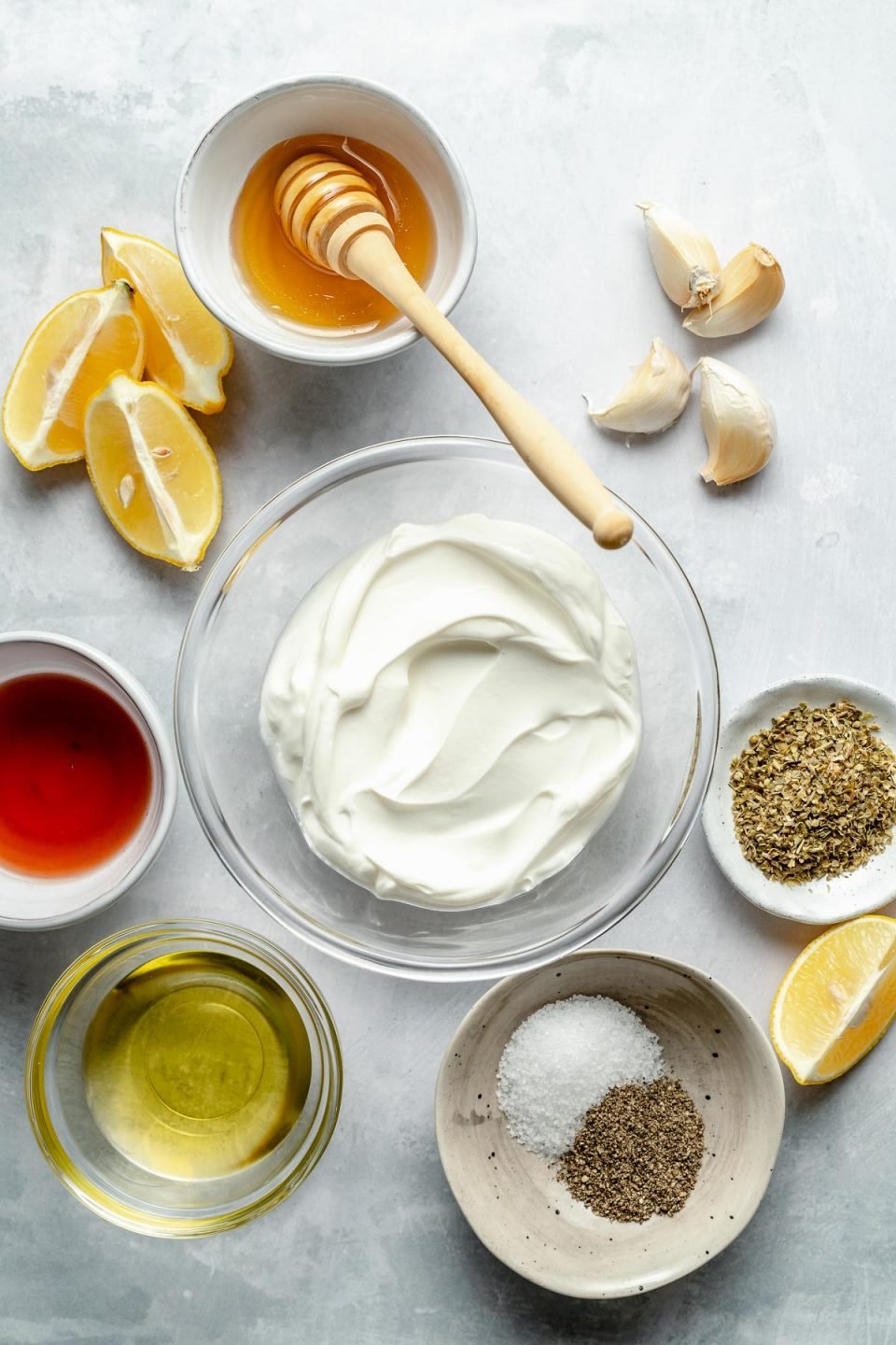 Greek marinade ingredients arranged on a light blue surface: greek yogurt, garlic, lemon, olive oil, red wine vinegar, honey, dried oregano, salt, & pepper.