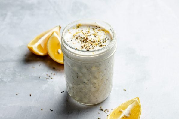 Greek marinade shown in a small ball jar, sitting atop a light blue surface with lemon wedges & dried herbs on the surface in the foreground & in the background.