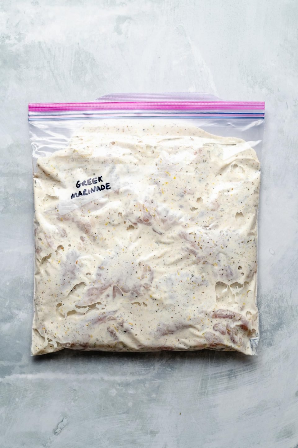 Chicken thighs in a Ziploc bag, marinating in Greek marinade. The bag sits atop a light blue surface. “Greek Marinade" is penned in the bag's memo area.