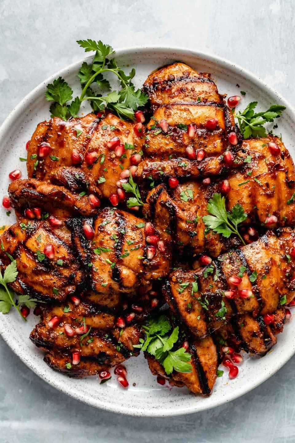 Grilled honey harissa marinated chicken thighs shown on a white speckled plate atop a light blue surface. The chicken is garnished with pomegranate arils & fresh cilantro leaves.
