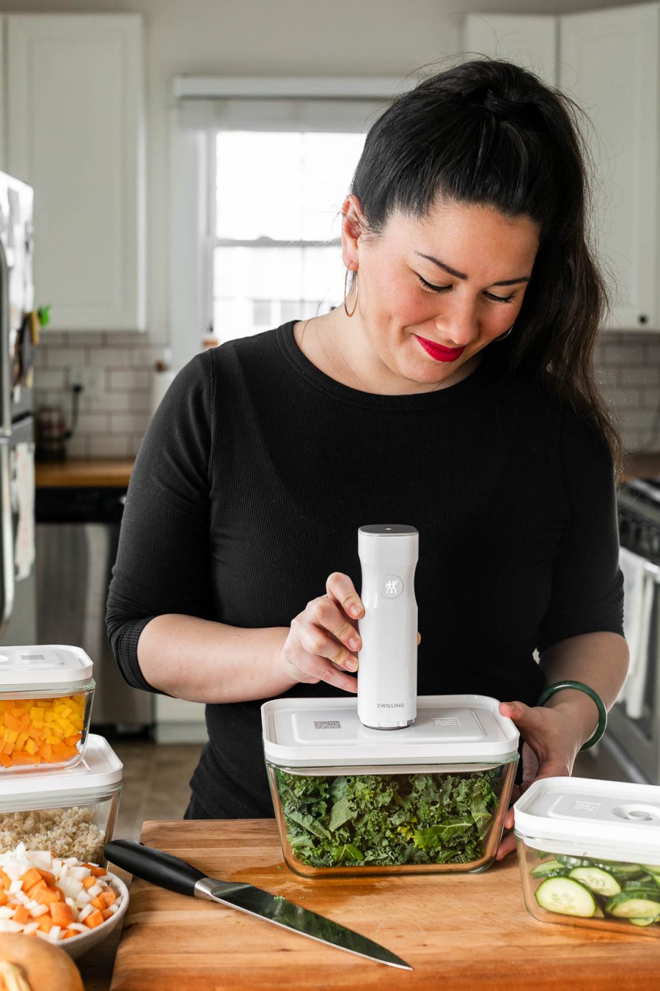 Jess standing at her kitchen counter vacuum sealing a Zwilling Fresh & Save container filled with shredded kale. The container sits atop a wood cutting board, next to a knife & some bowls filled with chopped veggies.