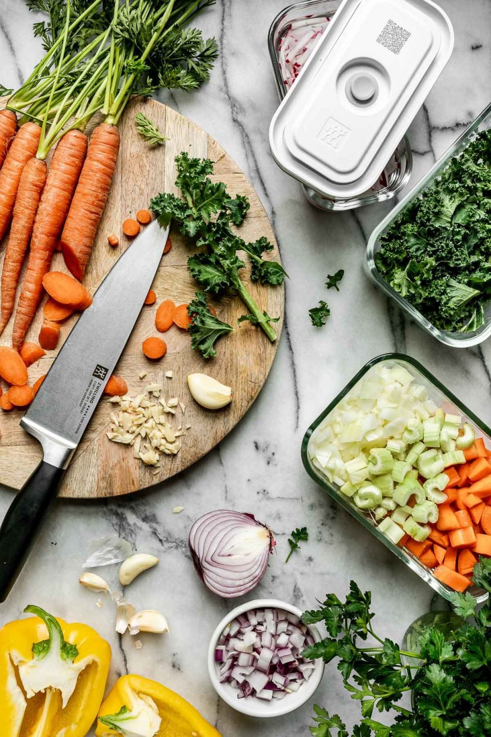 Mise en Place with Zwilling Fresh & Save. Zwilling Fresh & Save containers sit atop a white marble surface, filled with sliced radishes, shredded kale, & diced mirepoix. The containers are surrounded by fresh carrots, sliced garlic, diced onion, & halved bell pepper.