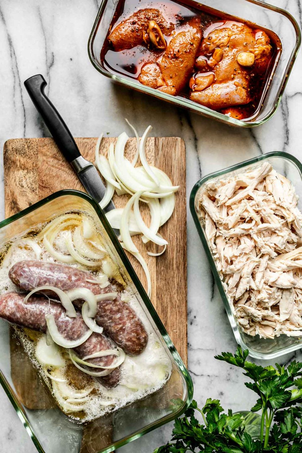 Mise en Place with Zwilling Fresh & Save. Zwilling Fresh & Save containers sit atop a white marble surface, filled with marinating chicken shawarma, shredded chicken breast, & marinating beer brats. The containers are surrounded by sliced onions & a bowl of fresh parsley.