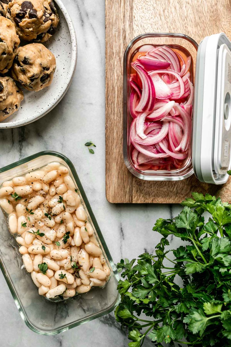 Mise en Place with Zwilling Fresh & Save. Zwilling Fresh & Save containers sit atop a white marble surface, filled with cooked white beans, pickled red onions. There is a plate of cookie dough & a bowl of fresh parsley on the surface, as well.