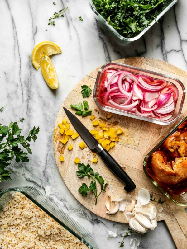 Mise en Place with Zwilling Fresh & Save. Zwilling Fresh & Save containers sit atop a white marble surface, filled with shredded kale, pickled red onions, marinating chicken shawarma, & brown rice. The containers are surrounded by lemon wedges, fresh thyme leaves, garlic, & a small wooden cutting board.