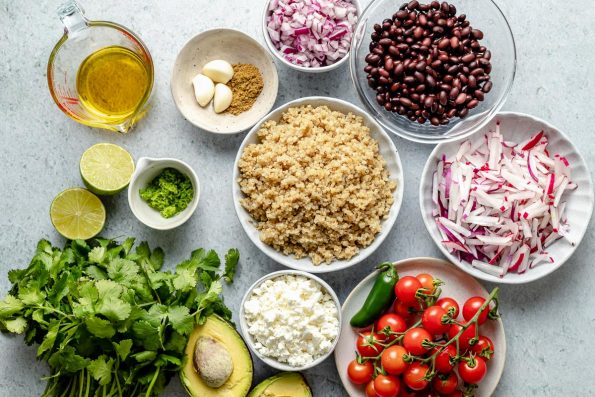Southwest quinoa salad ingredients arranged on a light blue surface – olive oil, limes, lime zest, cilantro, avocado, queso fresco, quinoa, garlic, cumin, red onion, black beans, radish, jalapeno, & cherry tomatoes.