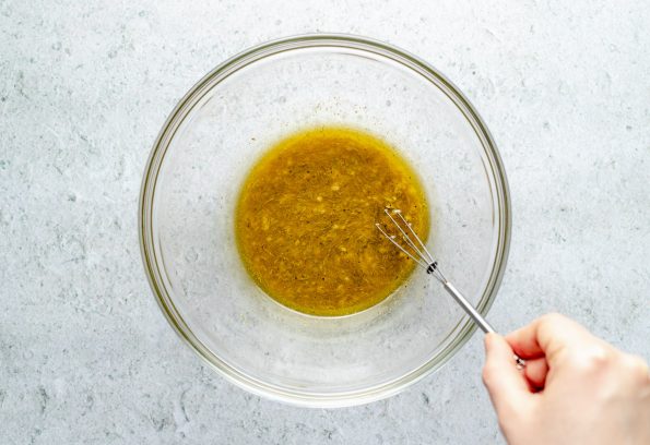 How to make Southwest Quinoa Salad, Step 1 – Cumin-Lime dressing in a large glass mixing bowl on a light blue surface, with a woman's hand shown whisking it together.