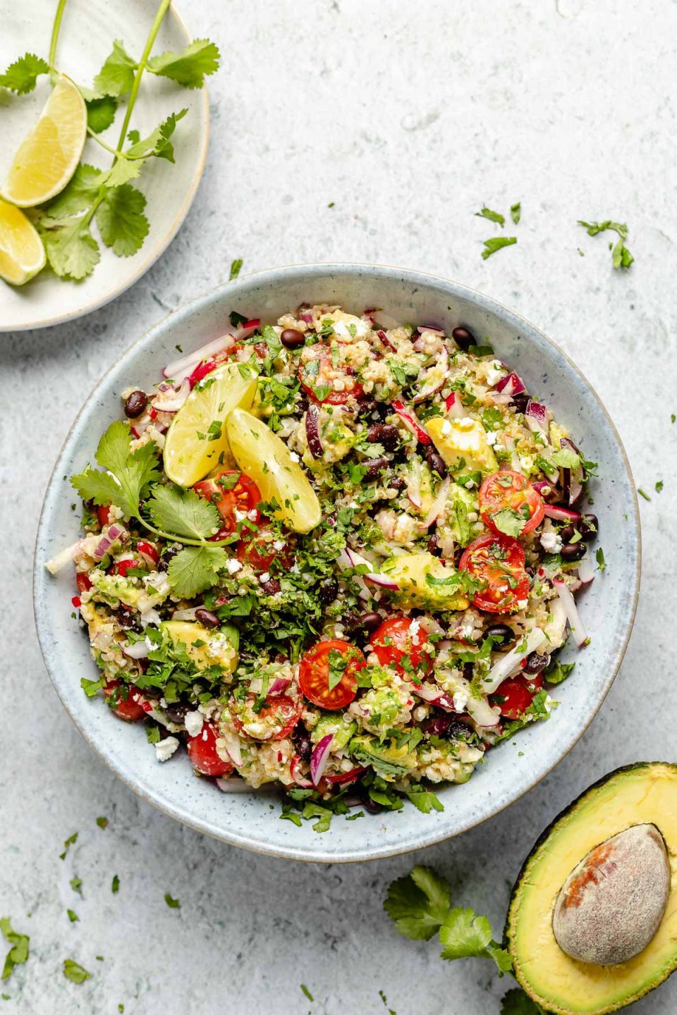 Southwest quinoa salad shown in light blue serving bowl, topped with fresh cilantro & lime wedges. The bowl sits atop a light blue surface, surrounded by chopped cilantro, lime wedges, & halved avocado.