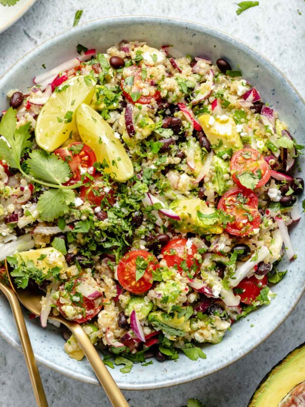 Close up of Southwest quinoa salad shown in light blue serving bowl, topped with fresh cilantro & lime wedges. Gold flatware is nestled into the quinoa salad for serving. The bowl sits atop a light blue surface, surrounded by chopped cilantro, lime wedges, & halved avocado.