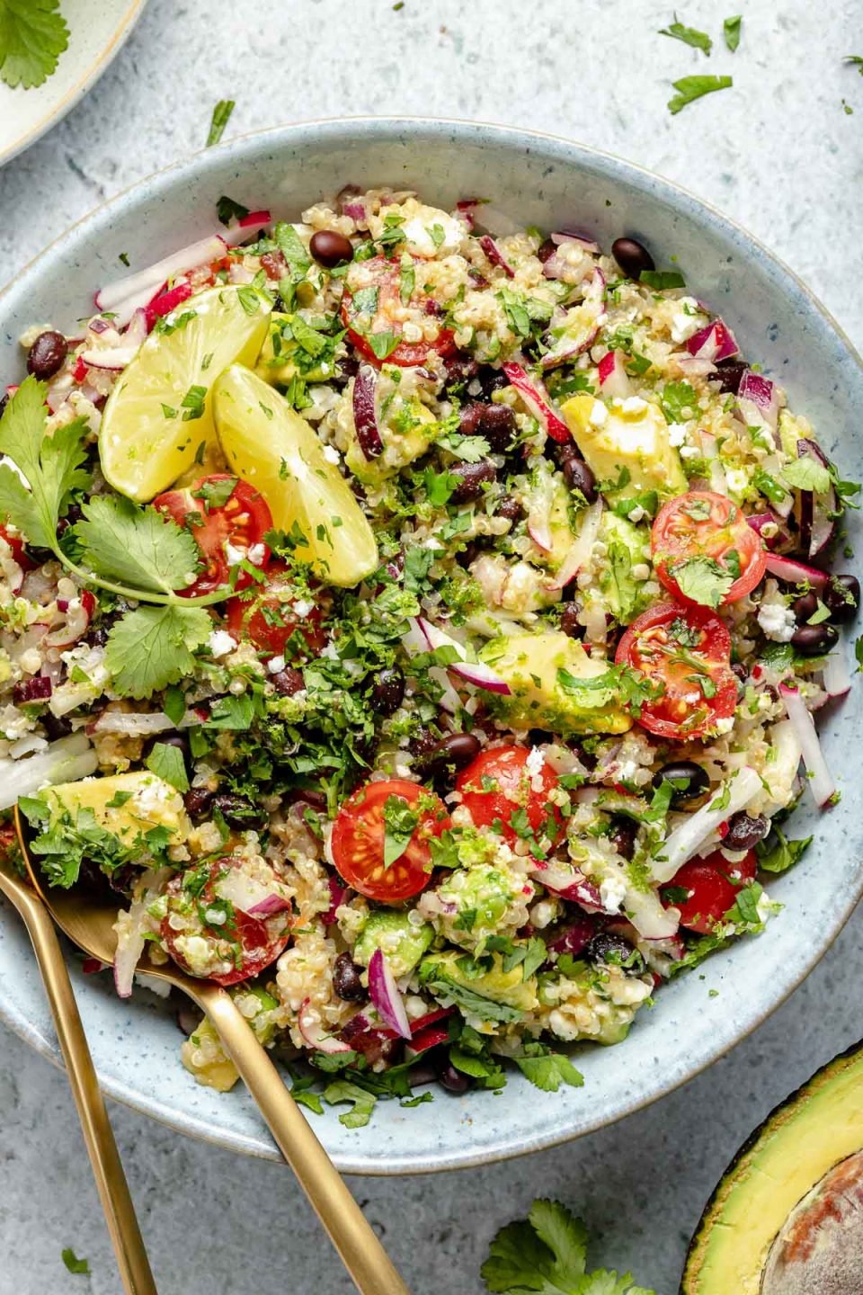 Close up of Southwest quinoa salad shown in light blue serving bowl, topped with fresh cilantro & lime wedges. Gold flatware is nestled into the quinoa salad for serving. The bowl sits atop a light blue surface, surrounded by chopped cilantro, lime wedges, & halved avocado.
