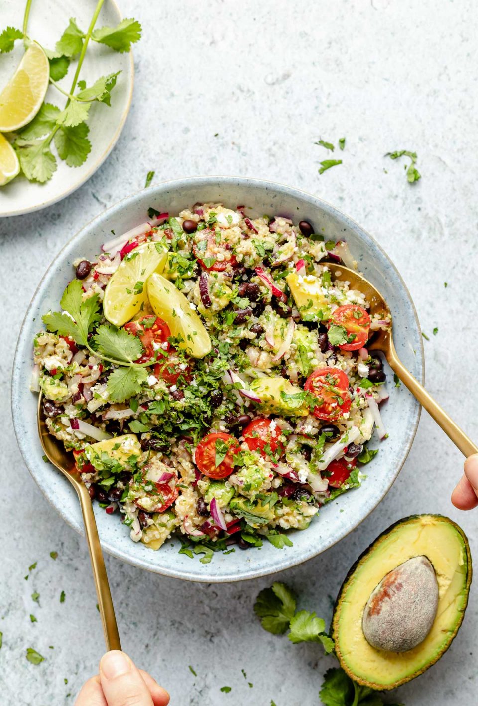 Close up of Southwest quinoa salad shown in light blue serving bowl, topped with fresh cilantro & lime wedges. A woman's hands are shown holding gold flatware, serving the salad. The bowl sits atop a light blue surface, surrounded by chopped cilantro, lime wedges, & halved avocado.